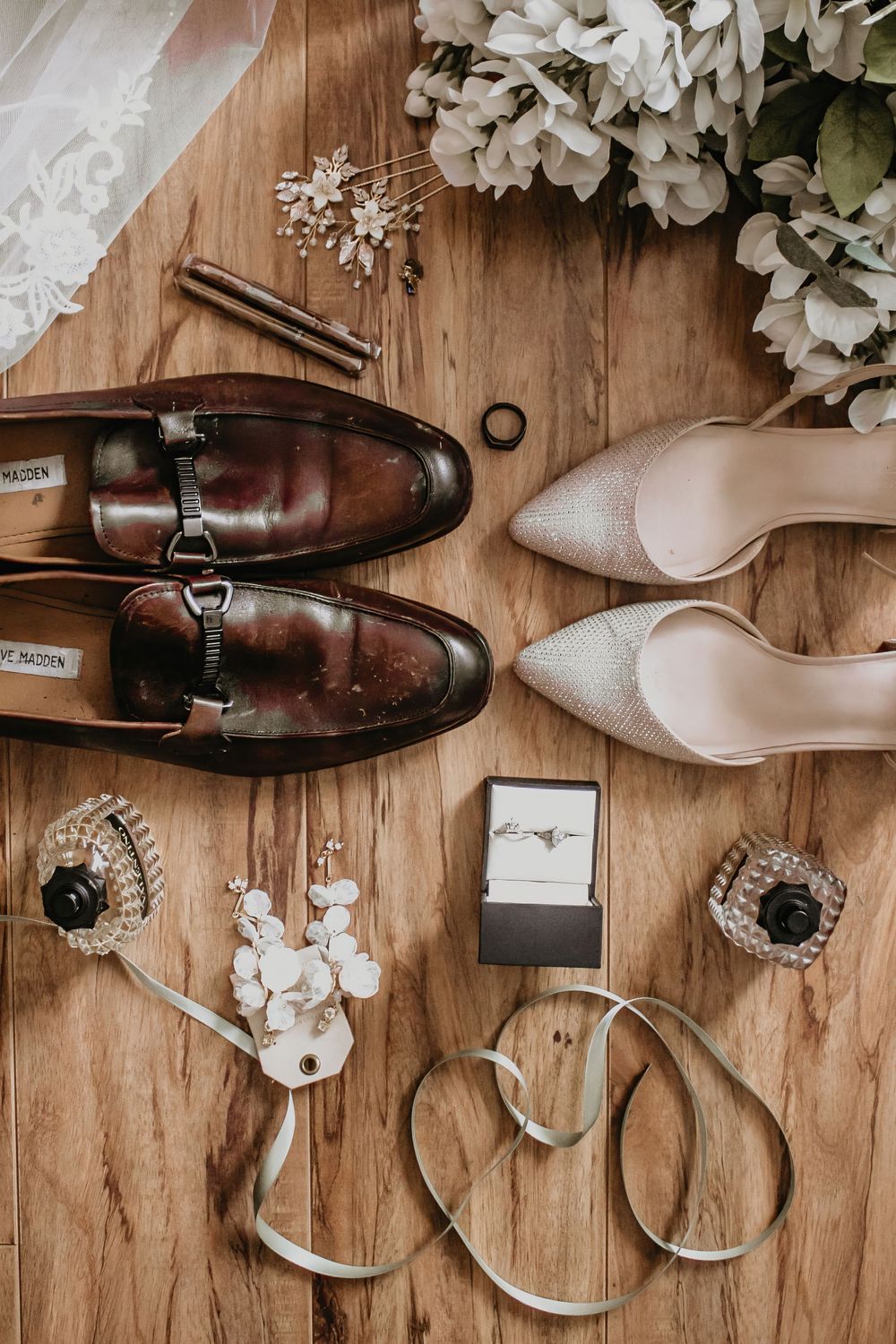 Flat lay of wedding details including leather loafers, heels, rings, bouquet, and accessories on wood.