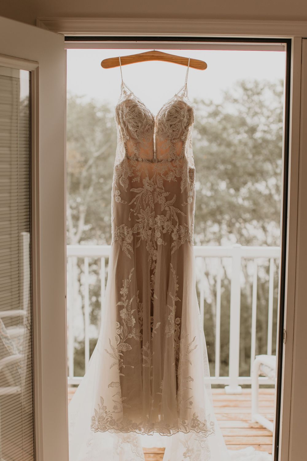 Elegant lace and tulle wedding gown hanging on wooden hanger in sunlit balcony doorway.