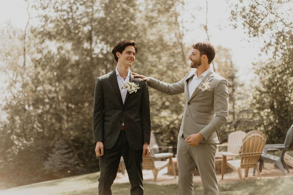 Two groomsmen in suits sharing a friendly moment outdoors with trees and wooden chairs in the background.