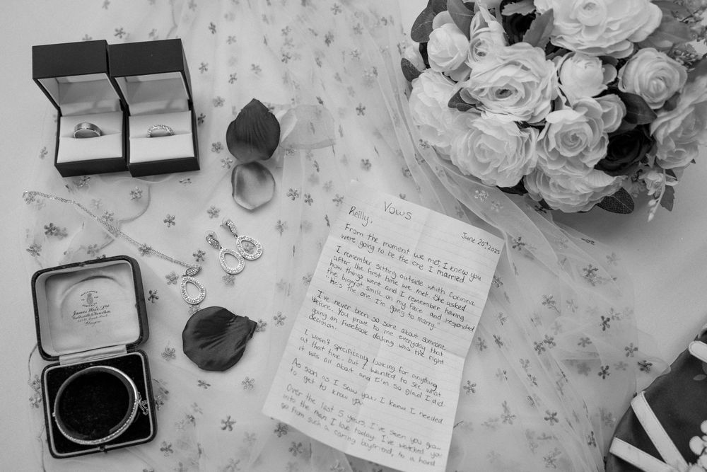 Flat lay of wedding rings, jewelry, handwritten letter, and white rose bouquet in black and white.