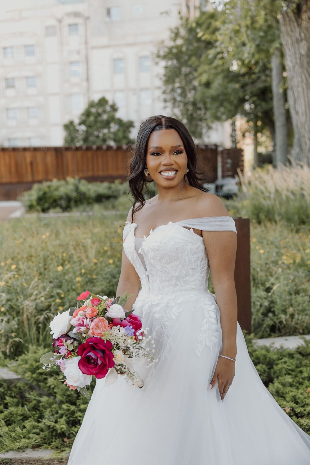 Smiling bride in off-shoulder lace wedding gown holds vibrant pink and coral bouquet outdoors.