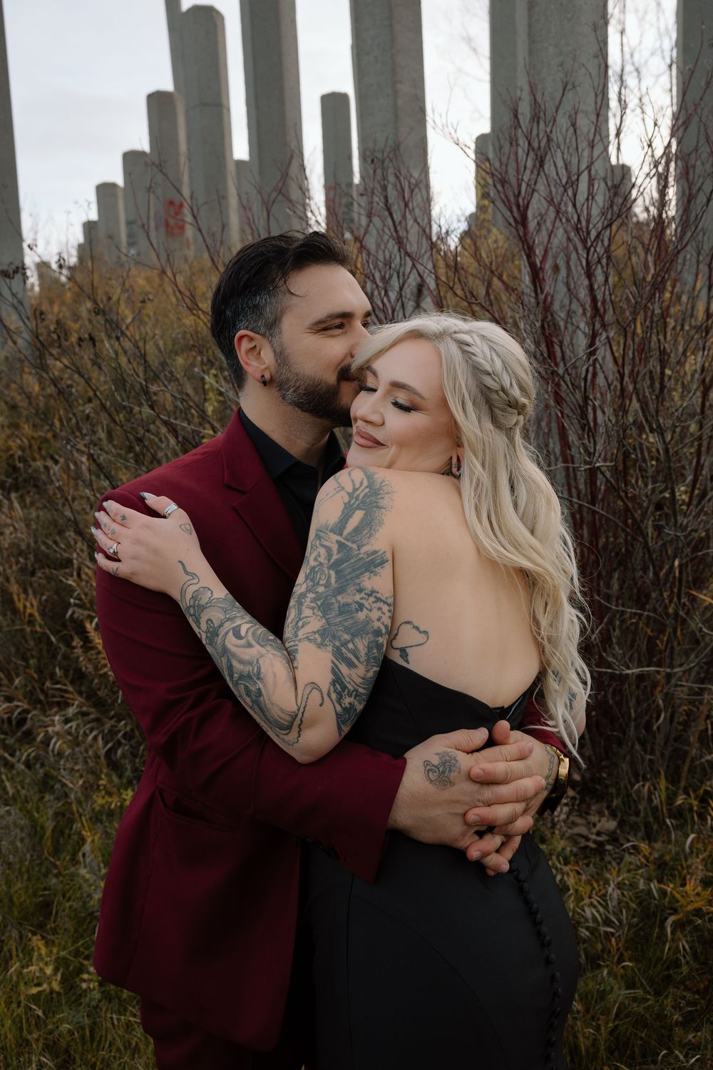 Tattooed couple embracing in an outdoor autumn setting with concrete pillars, man in burgundy suit kissing her cheek.