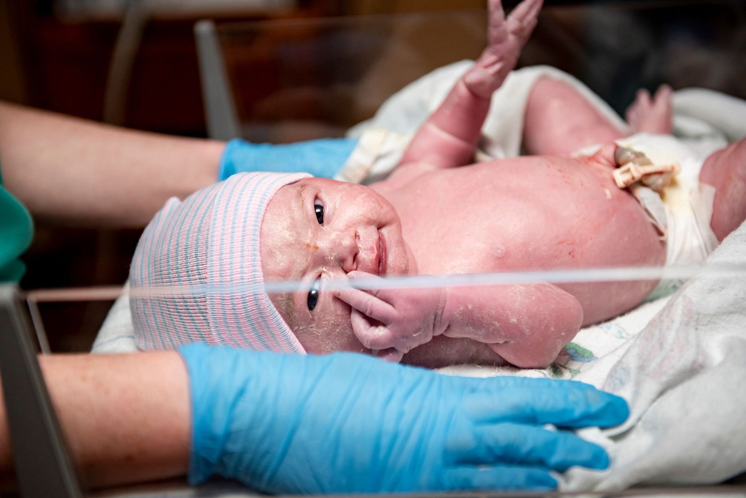A newborn baby smiles while being examined by medical staff wearing blue gloves.
