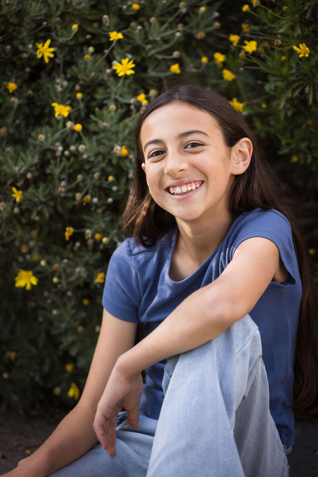 Smiling young girl in blue t-shirt sitting outdoors in front of green bushes with yellow flowers.
