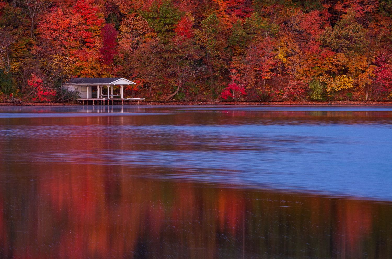 Autumn lake reflection with vibrant red foliage and a boathouse mirrored in calm water.