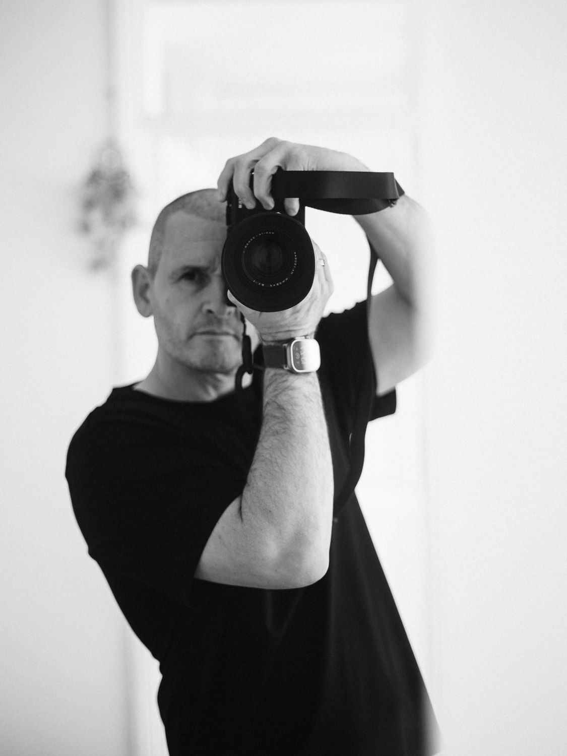 Bald man in black t-shirt taking a self-portrait with a DSLR camera in a bright white room.