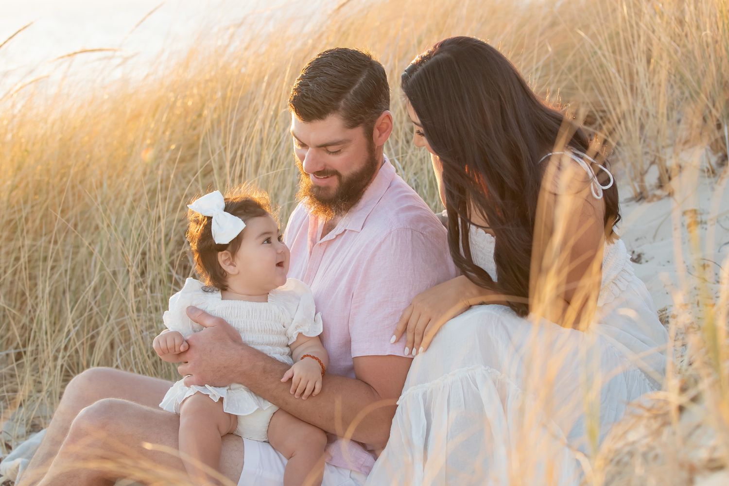 Family sitting in golden beach grass at sunset, parents smiling at baby girl wearing white dress and bow.