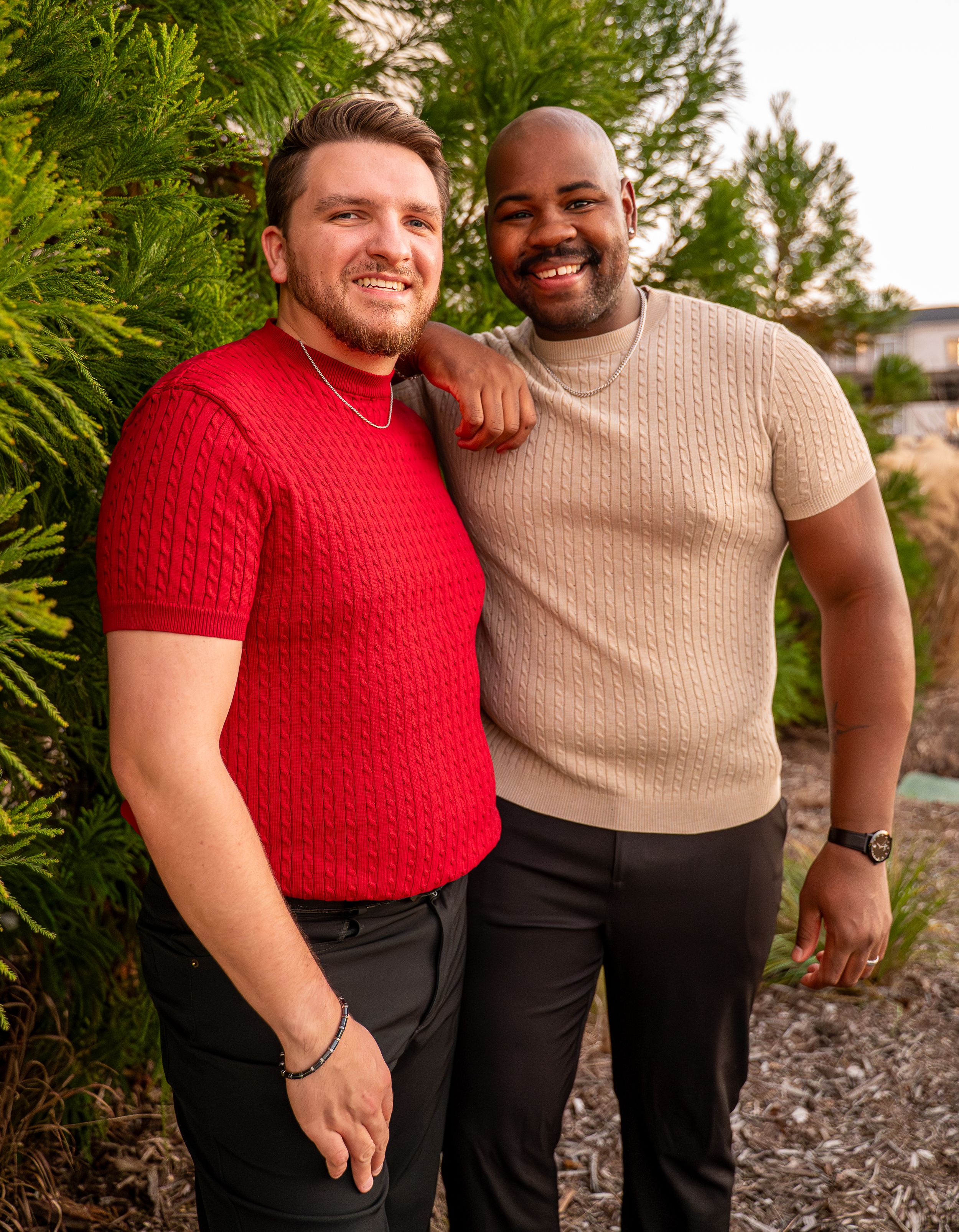 Two smiling men posing together outdoors near green trees at dusk.