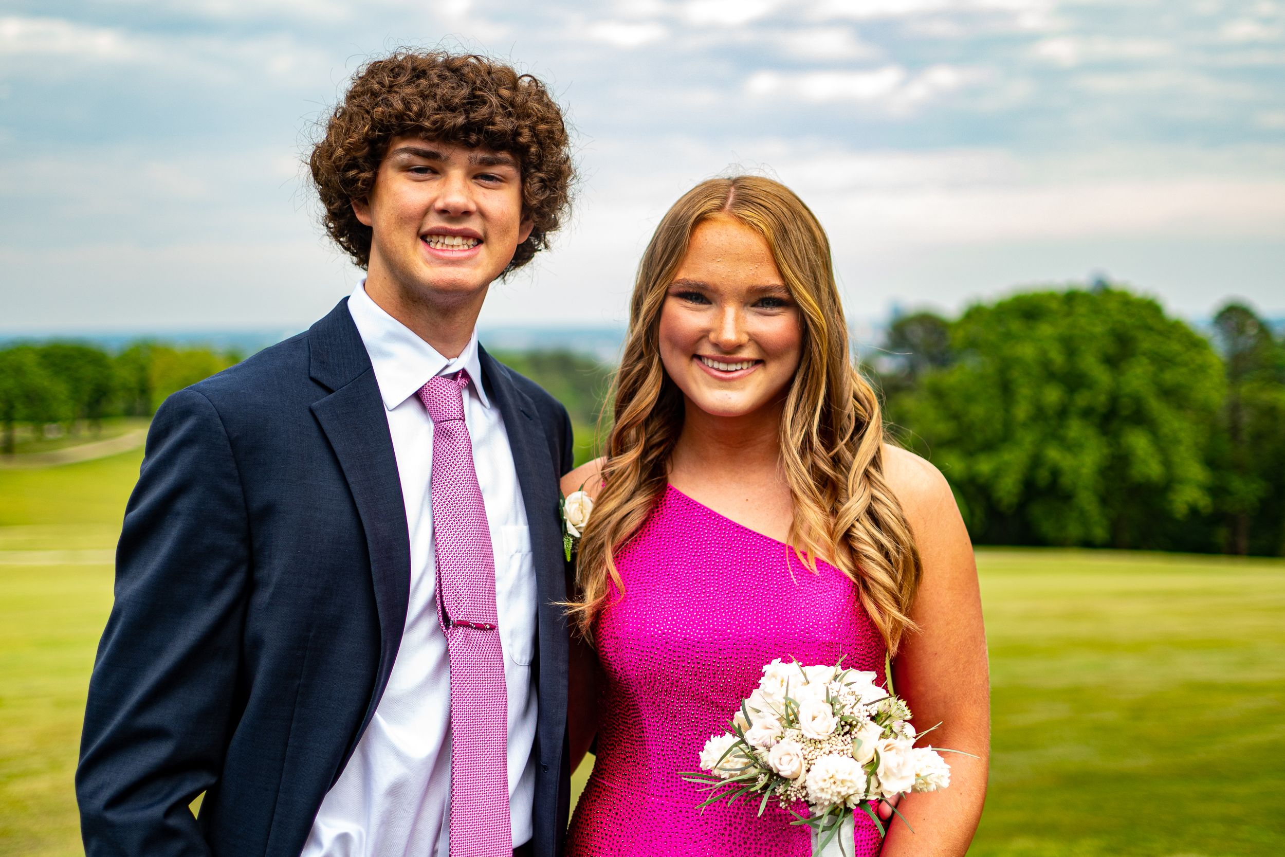 Smiling prom couple standing on golf course, young man in navy suit and girl in hot pink dress holding white bouquet.