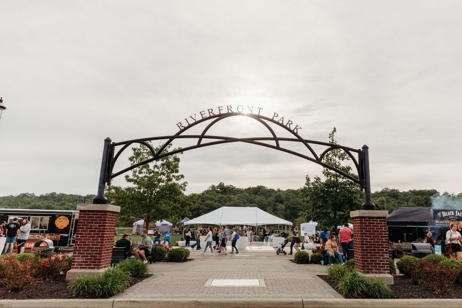 Riverfront Park entrance arch with brick pillars, outdoor event tents, and visitors on a cloudy day.