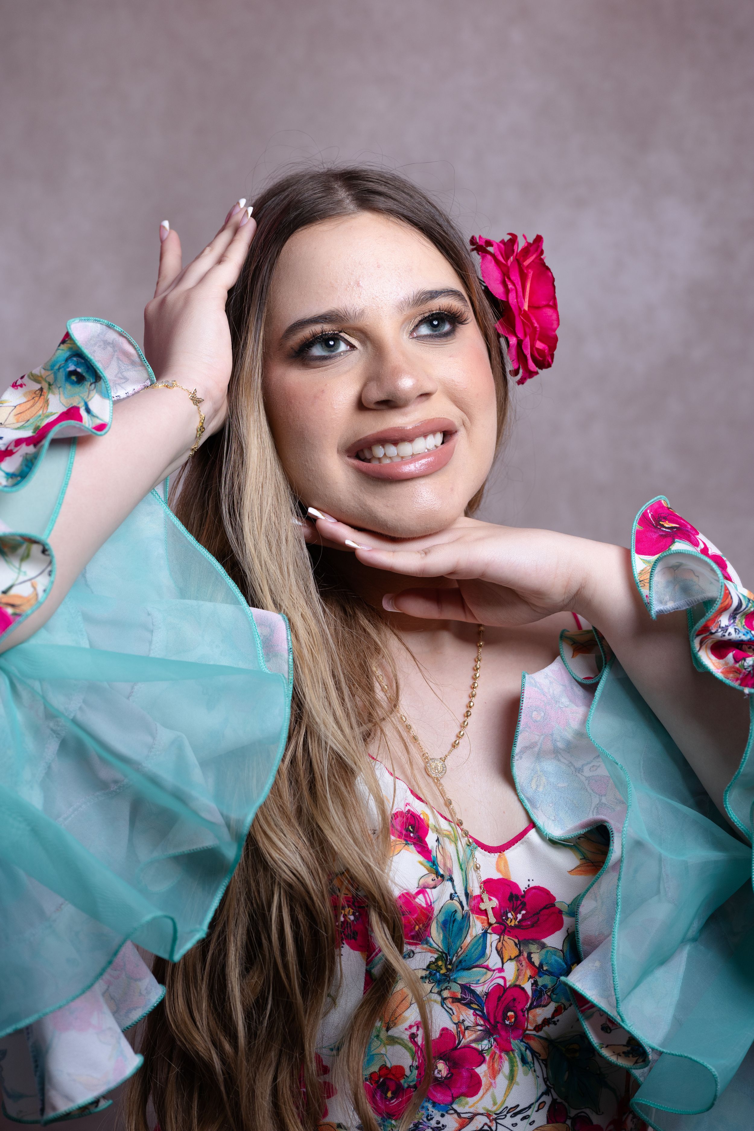 Young woman in floral dress with teal ruffles and pink flower hair accessory smiling in studio portrait.
