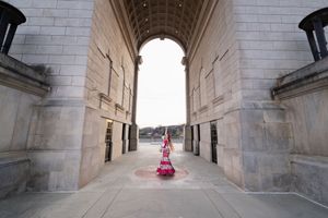 Woman in vibrant red and white dress standing under grand stone archway of historic monument.