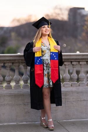 Smiling female graduate in black cap and gown wearing Venezuelan flag stole, posing outdoors at sunset.