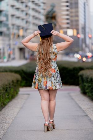 Young woman in floral dress and heels holding graduation cap, standing on urban pathway from behind.