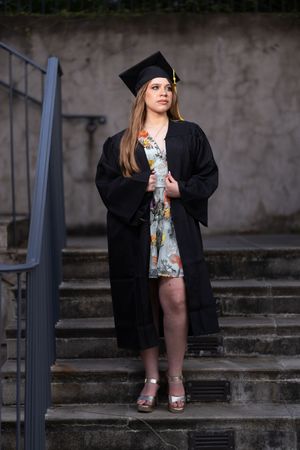 Female graduate in black cap and gown standing on stone steps holding diploma, floral dress visible.