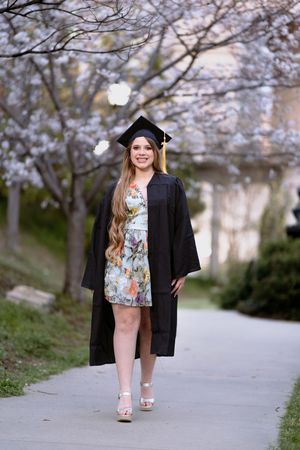 Smiling graduate in black cap and gown posing under cherry blossom trees on pathway.