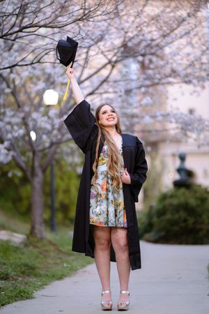 Smiling female graduate in black gown raises cap among cherry blossoms on a scenic campus pathway.