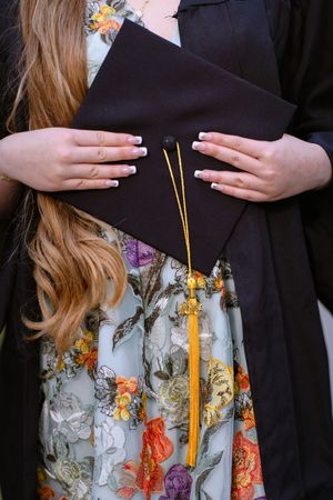 Close-up of graduate holding black mortarboard cap with gold tassel, wearing floral dress and black gown.