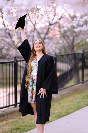 Joyful female graduate in black cap and gown tossing hat under blooming cherry blossom trees.
