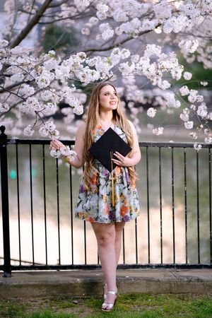 Young woman in floral dress holding graduation cap under blooming cherry blossom tree.