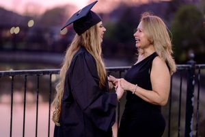 Graduate in cap and gown smiles with proud woman near waterfront at dusk.