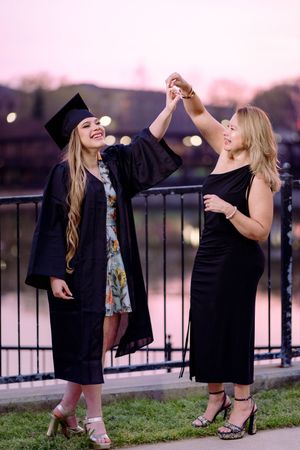 Two women celebrate graduation at dusk, one in cap and gown, holding hands raised near a railing.