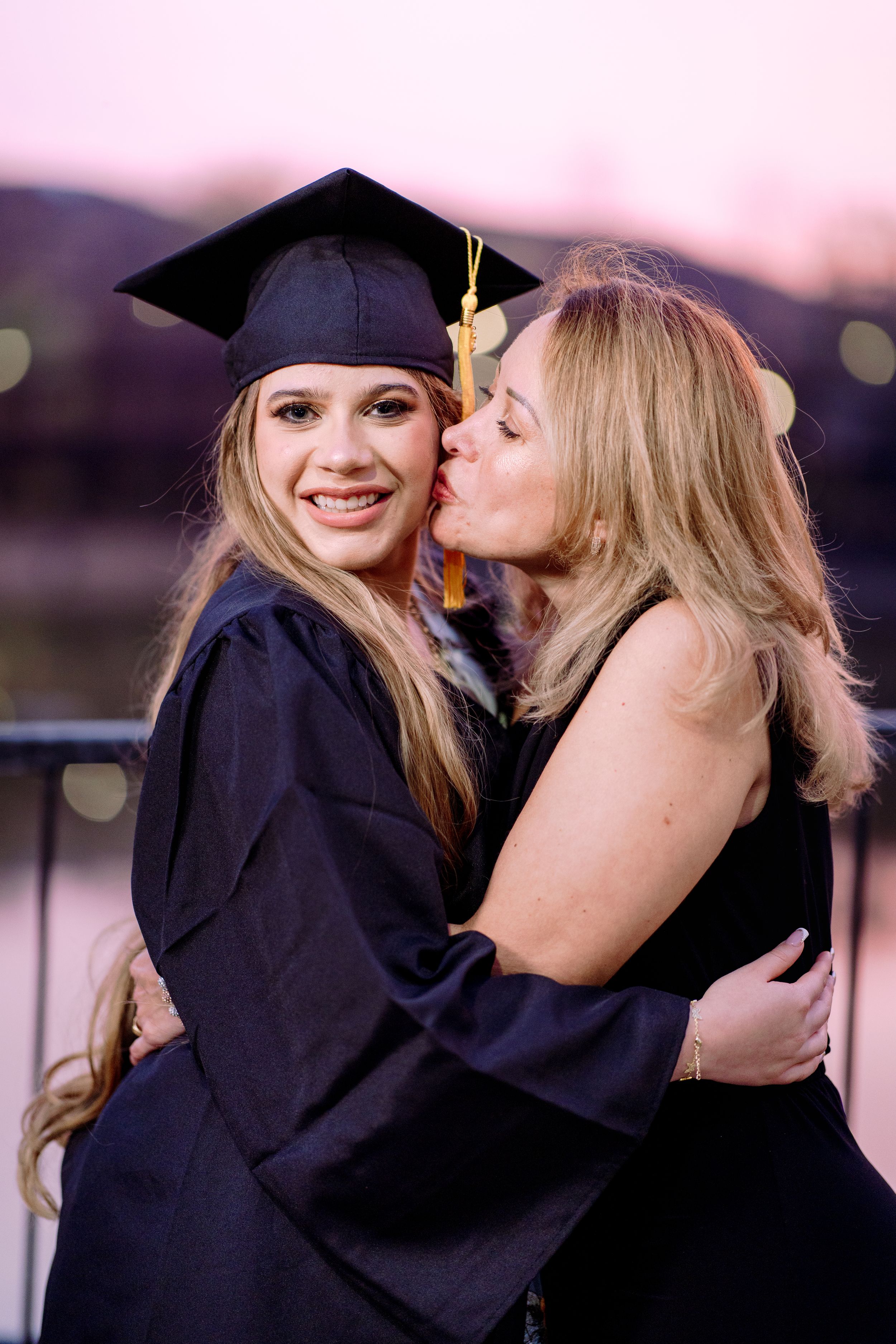 A smiling graduate in cap and gown receives a kiss on the cheek from a proud woman at a twilight outdoor ceremony.
