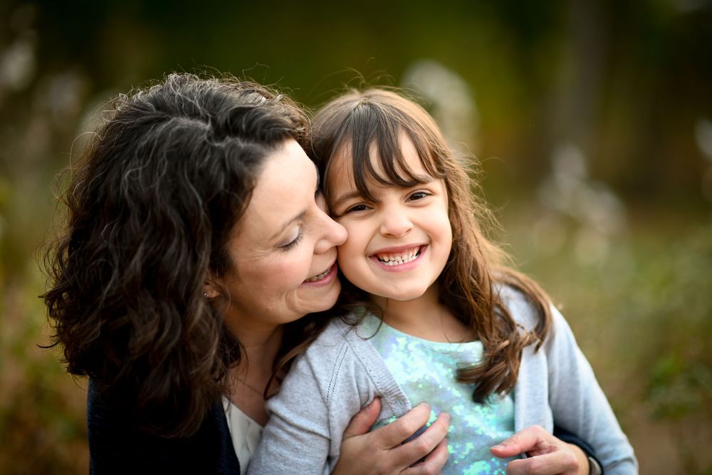 A mother hugging and kissing her smiling young daughter outdoors with a blurred green background.