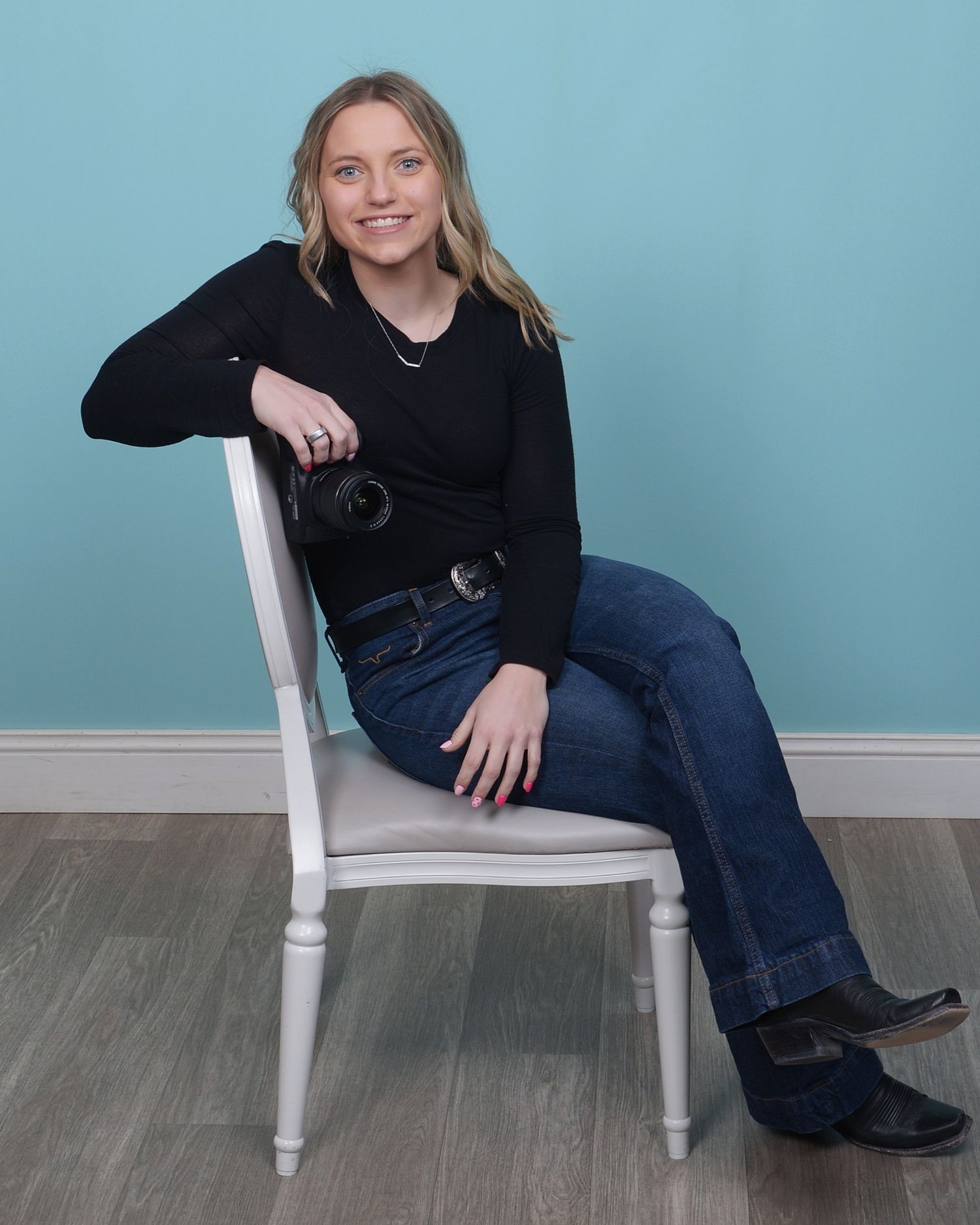 Portrait of someone in a black sweater and jeans sitting casually on a white chair against a light blue backdrop.