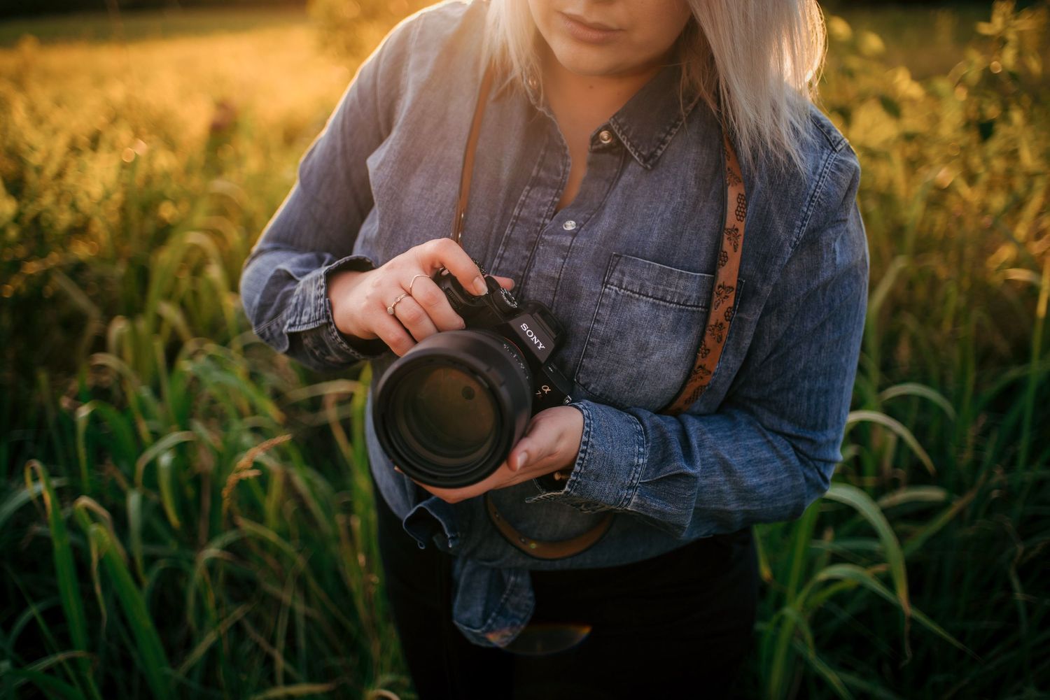 Photographer holding professional camera wearing denim shirt standing in golden wheat field at sunset.