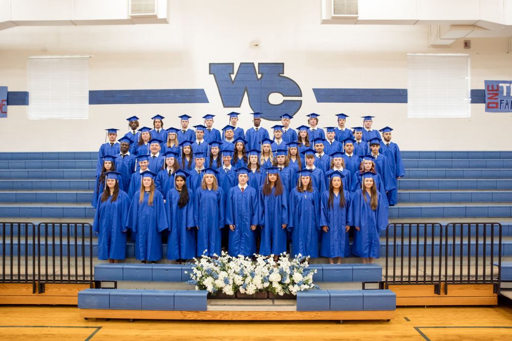 A group in blue graduation gowns poses on bleachers in a gymnasium with a floral arrangement in the foreground.