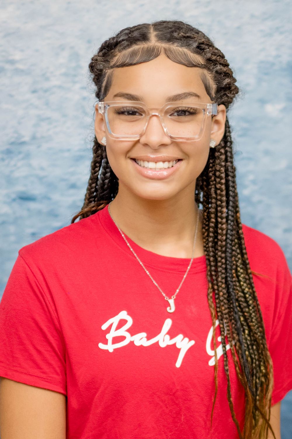 A smiling person in a red shirt with 'Baby' text wearing clear glasses and styled braids against a blue background.