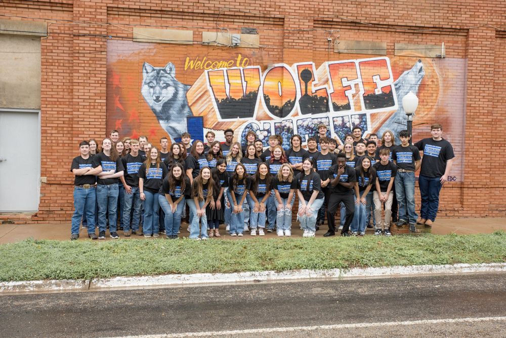 A large group in matching t-shirts poses together in front of a colorful wolf mural painted on a brick wall.
