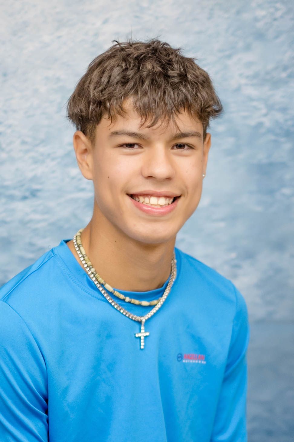 Young person in bright blue shirt with cross necklaces smiling against blue mottled backdrop.