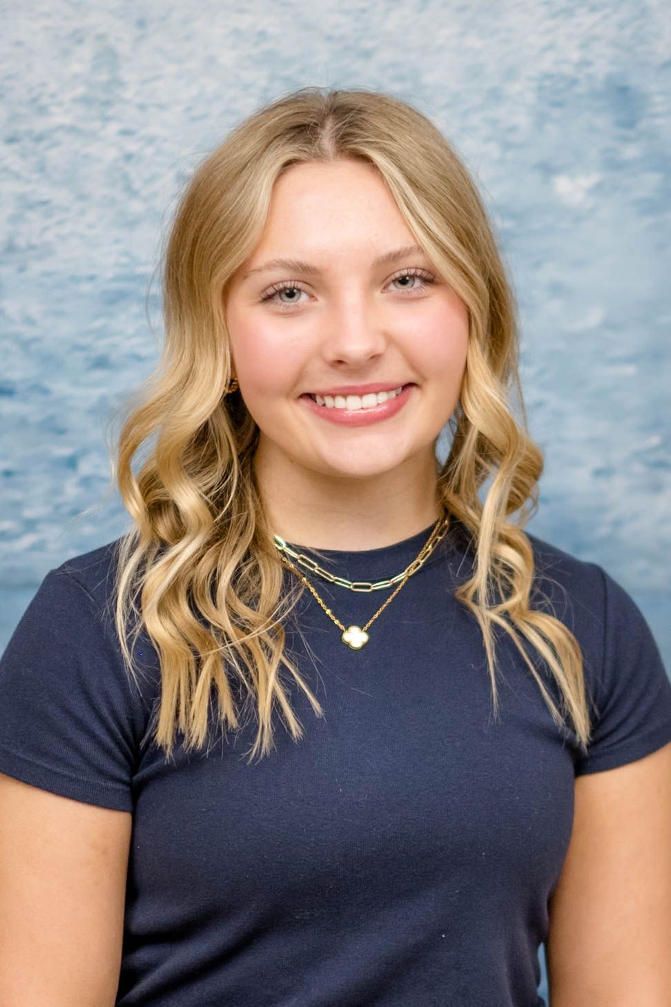 Portrait against blue textured background featuring blonde wavy hair, navy t-shirt and layered gold necklaces.