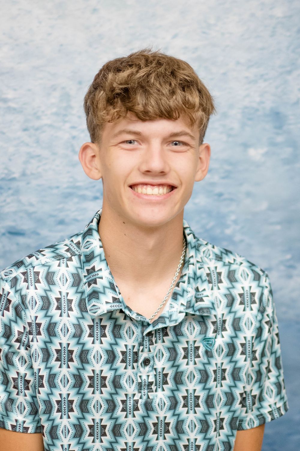 Young person in patterned short sleeve button-up shirt smiling against blue backdrop.
