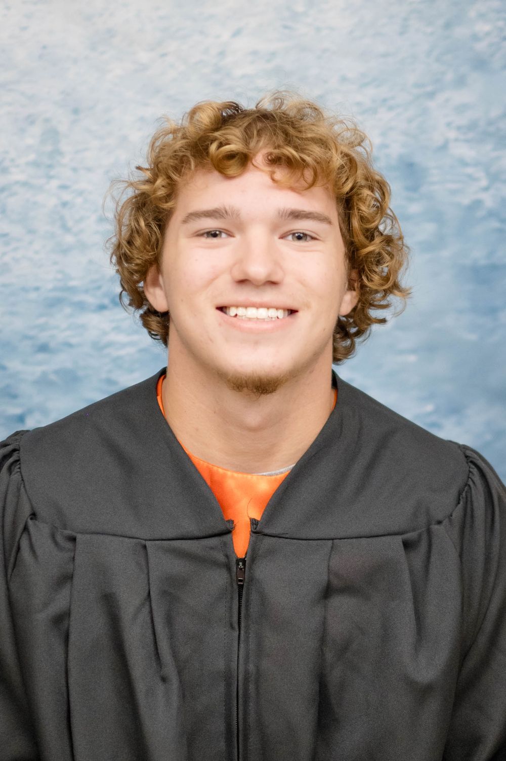 Graduate in black academic gown and orange shirt smiles against blue backdrop for portrait photo.