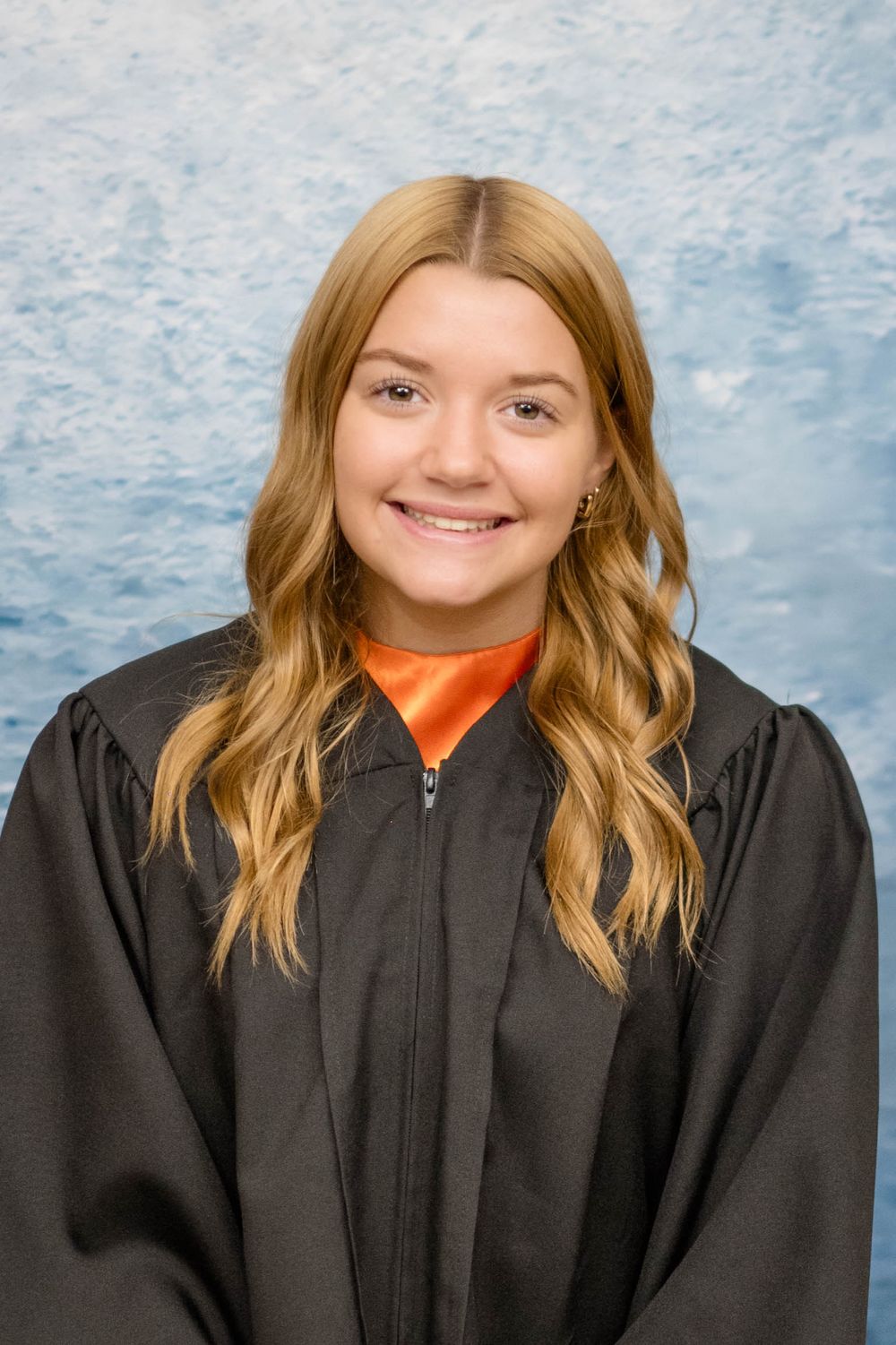 A graduate wearing a black academic gown and orange top poses against a mottled blue backdrop for a portrait.