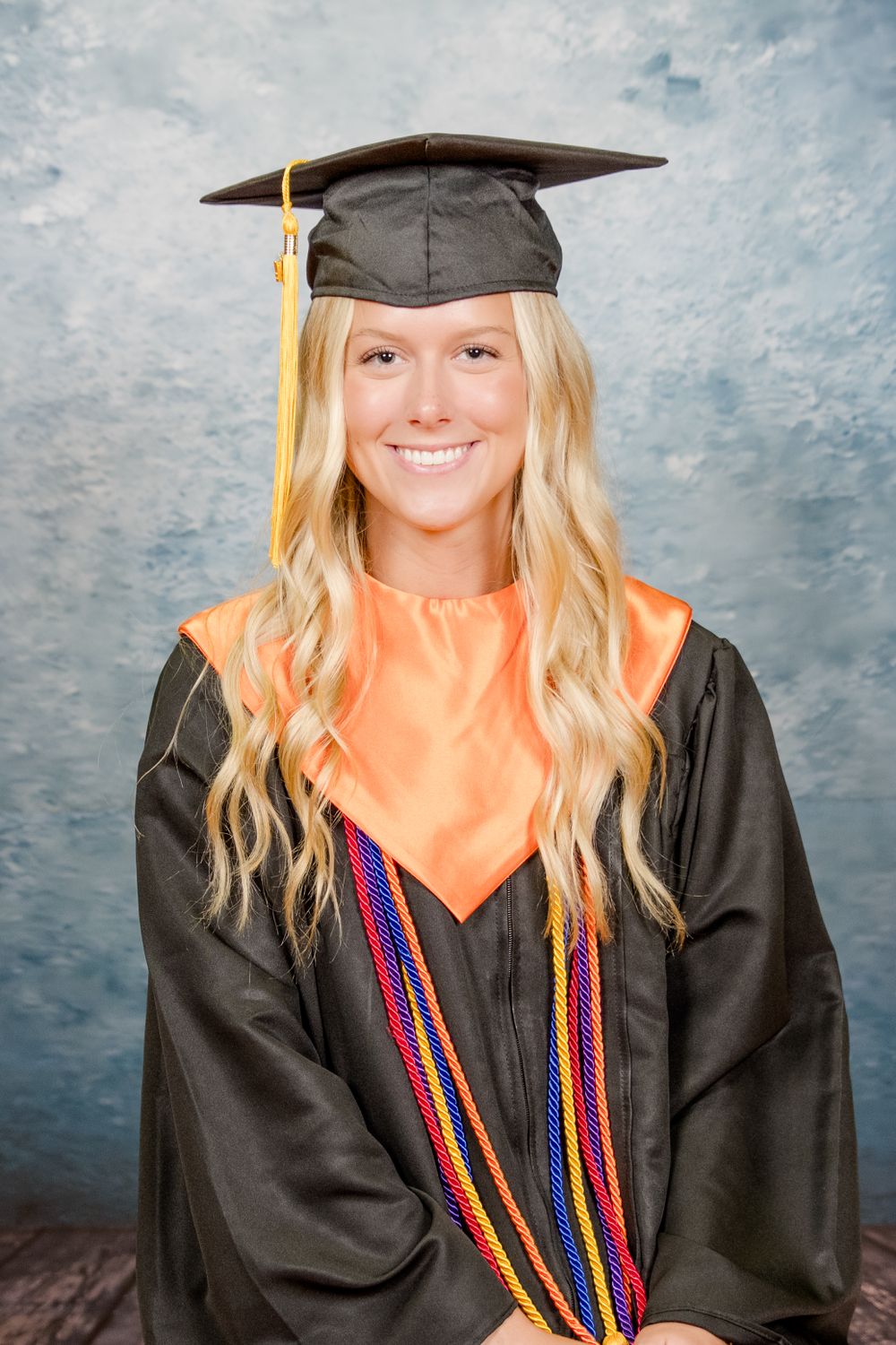 Graduate wearing black cap and gown with colorful honor cords and orange blouse smiles in front of blue backdrop.