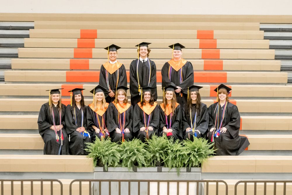 A group of graduates in black caps and gowns pose on concrete steps with orange accents and greenery.