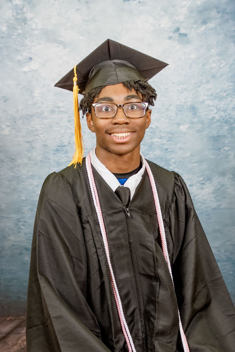 A graduate in traditional black cap and gown with honor cords smiles at the camera for their formal portrait.