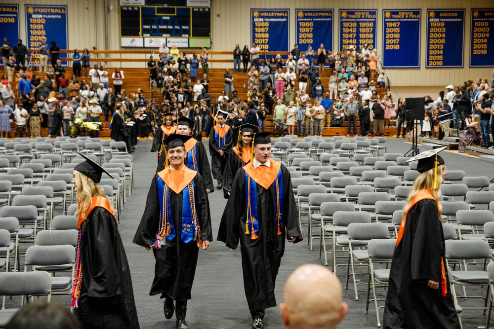 Graduates in black and blue academic regalia walk between rows of empty chairs during an indoor commencement ceremony.