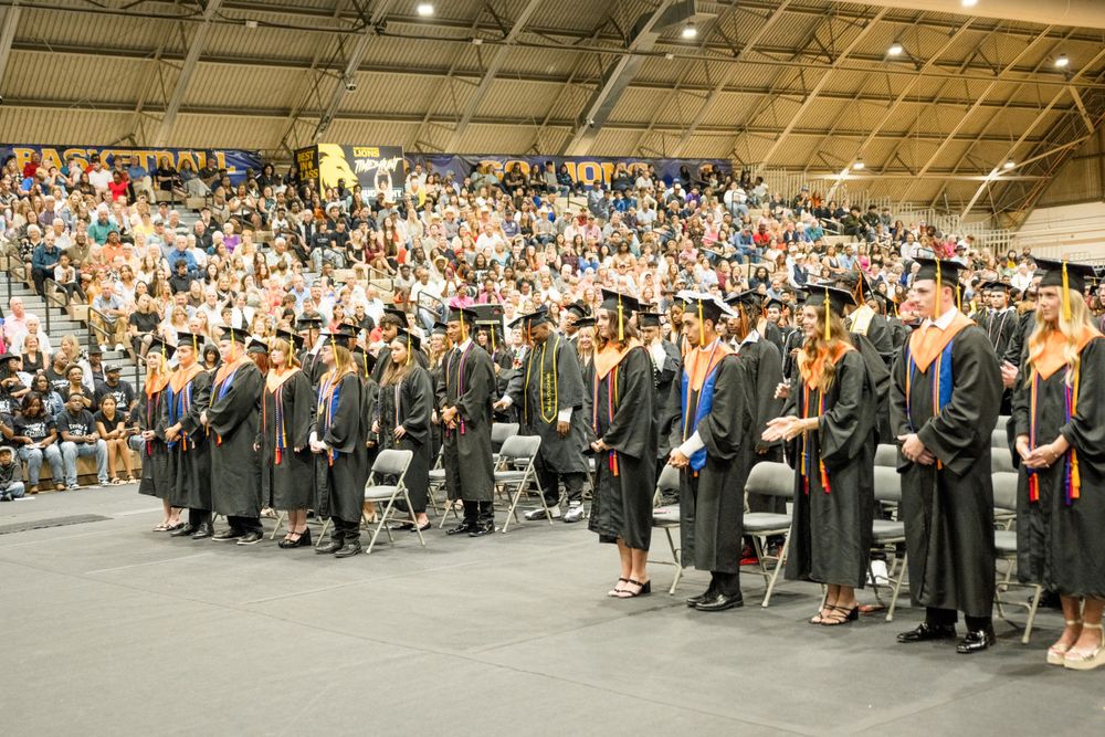 A large group of graduates in black caps and gowns stand during a commencement ceremony in an indoor arena.