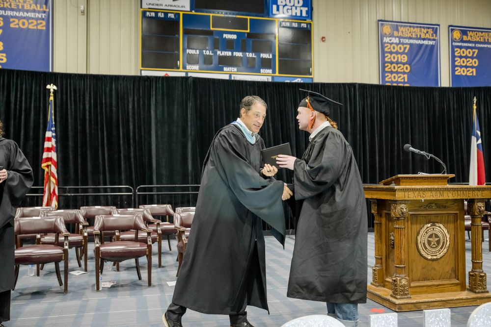 Graduates in formal academic regalia shake hands during a commencement ceremony on stage with flags and podium.