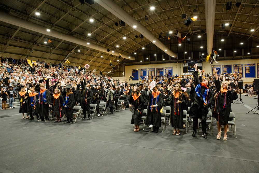 Graduates stand in a large indoor venue during a commencement ceremony with bright overhead lighting and filled bleachers.