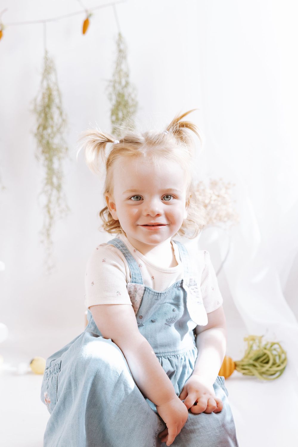 Small child wearing light blue overalls and white shirt sits on white backdrop with whimsical spring decor.