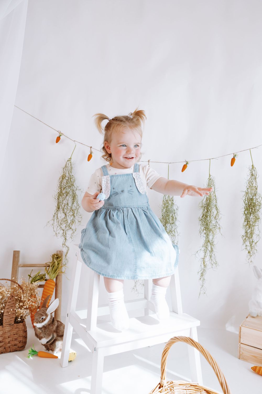A child in a blue dress stands on a white chair with spring decorations, Easter baskets, and fairy lights in backdrop.