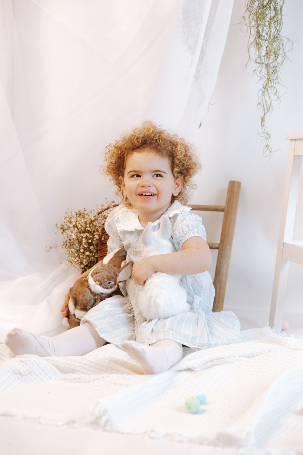 Child in white dress sitting on bed with curly hair while holding stuffed toy bunny in bright sunlit room.