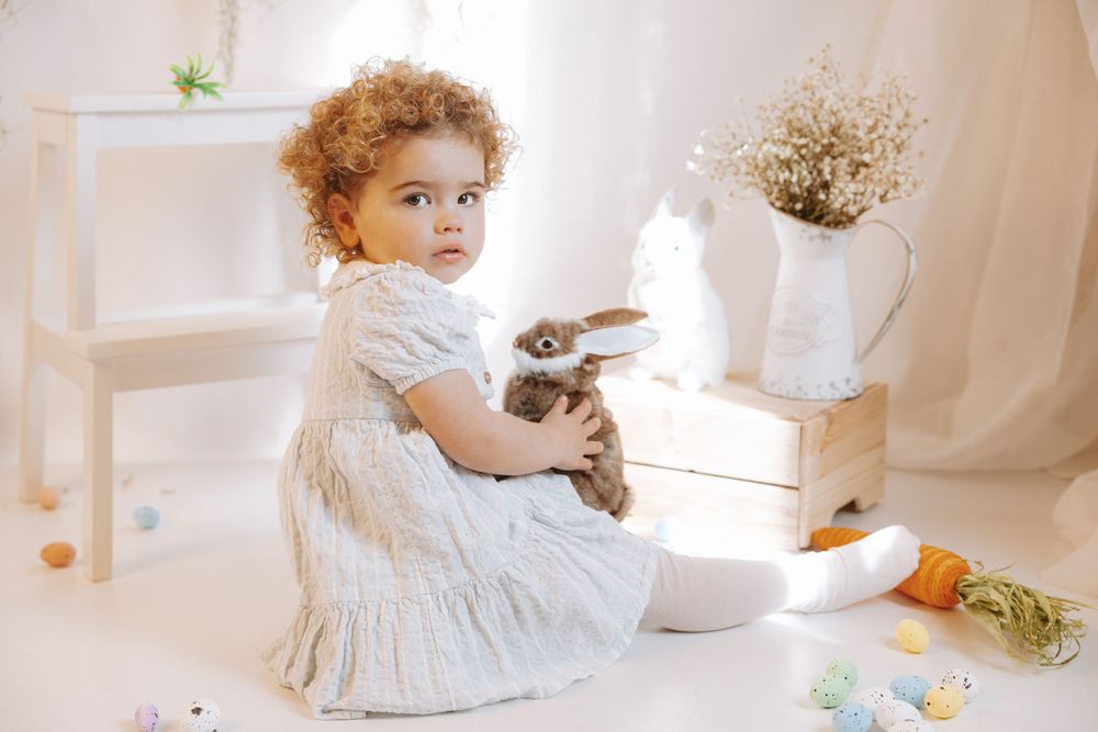 A small child in a white dress sits on the floor with a bunny rabbit in a bright, minimalist setting with dried flowers.