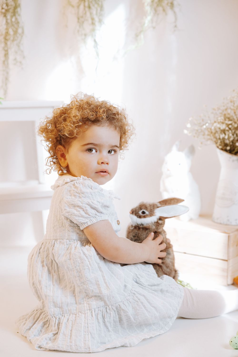 A child in a white linen dress sits with a plush bunny toy in a bright, airy room with neutral decor.